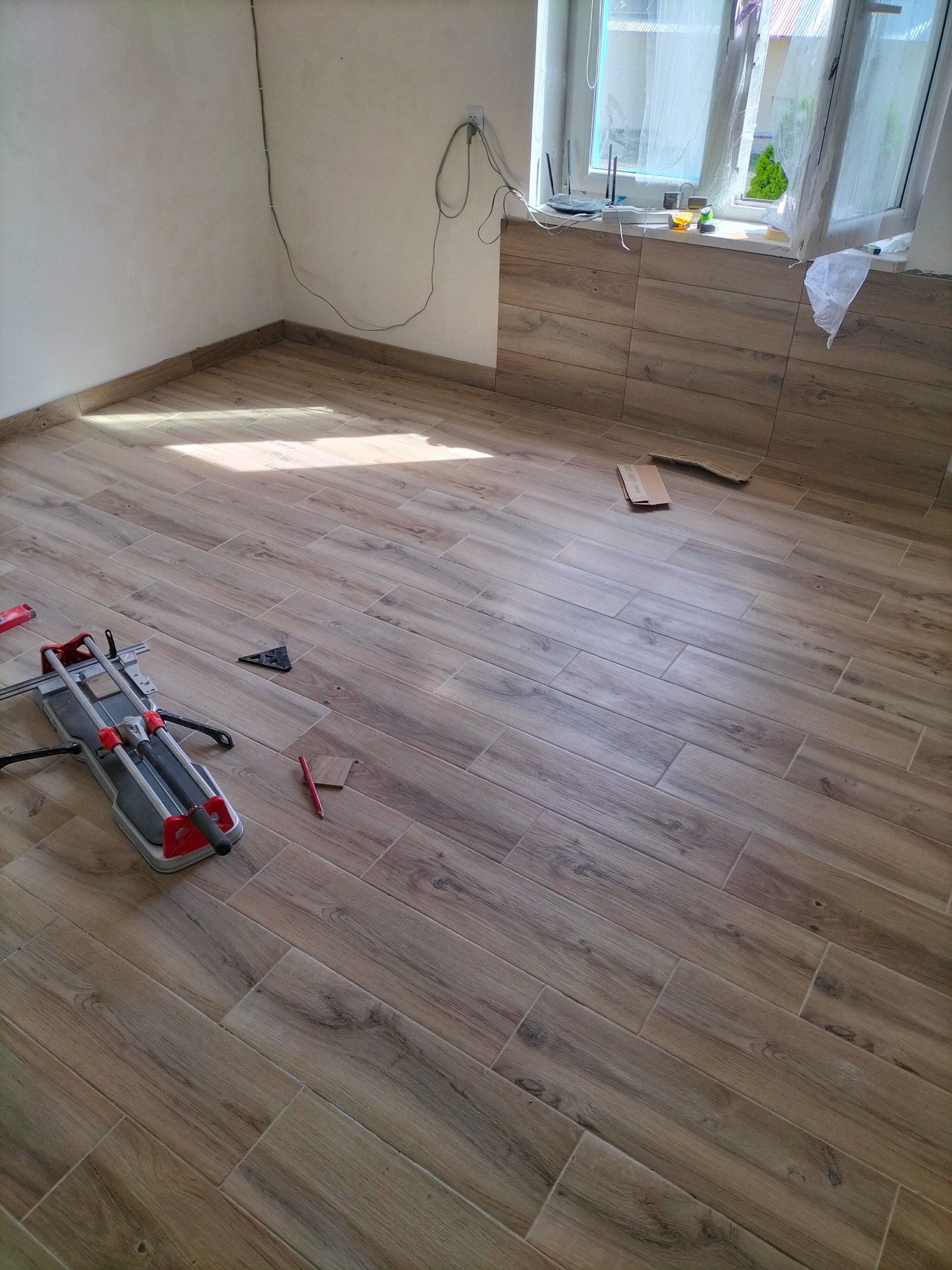 Układanie płytek podłogowych imitujących drewno w pomieszczeniu; widoczna przecinarka do płytek, poziomica i ołówek.