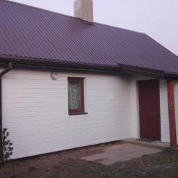 Yans - Dom jednorodzinny z białą elewacją panelową, ciemnobrązowym dachem z blachy trapezowej i brązowymi rynnami, widoczny z zewnątrz w pochmurny dzień.