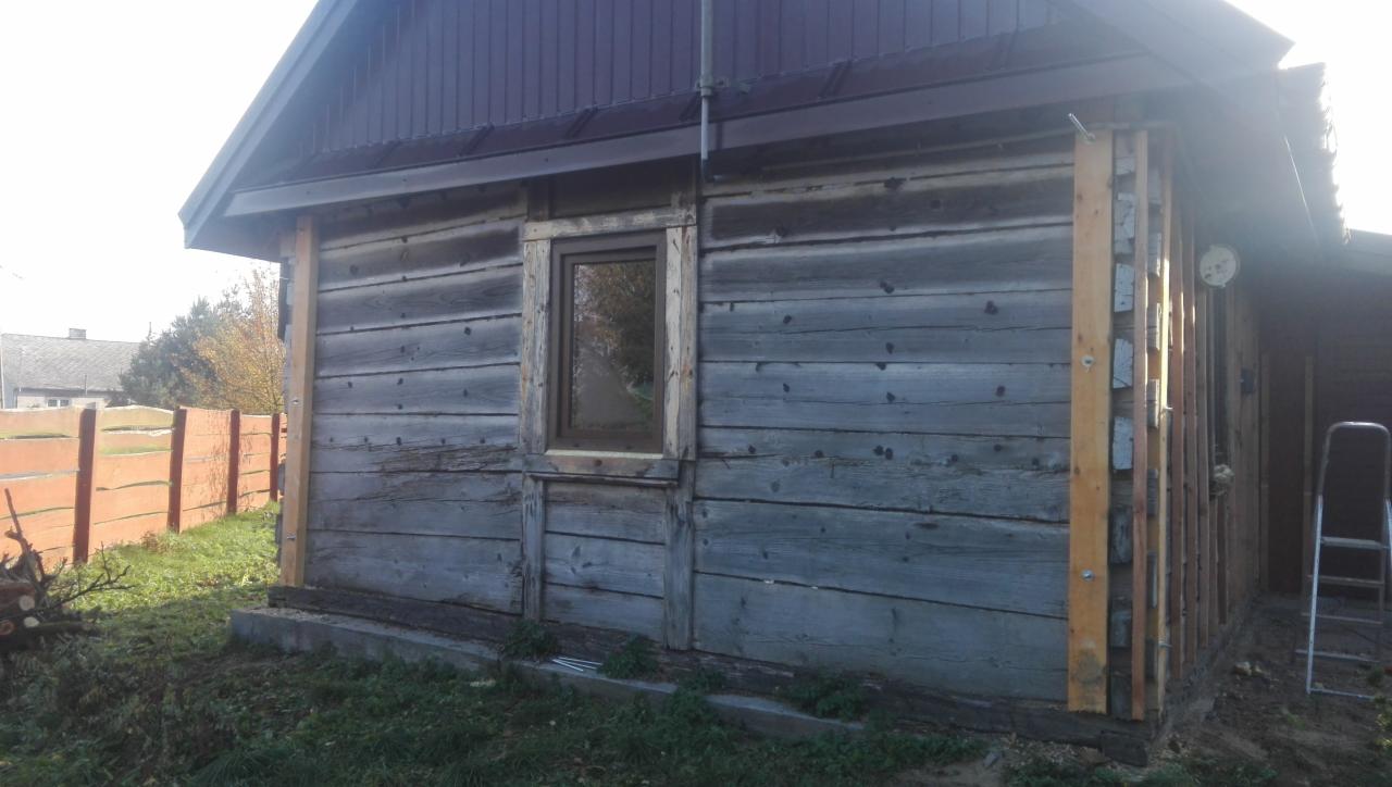 Drewniany budynek w trakcie renowacji, z widocznymi elementami konstrukcyjnymi i nowym oknem, drewniane deski na elewacji, fragment ogrodzenia w tle.