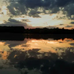 Spokojna tafla jeziora odbija dramatyczne chmury oświetlone zachodzącym słońcem, tworząc symetryczny krajobraz z ciemnym pasem lasu na horyzoncie.