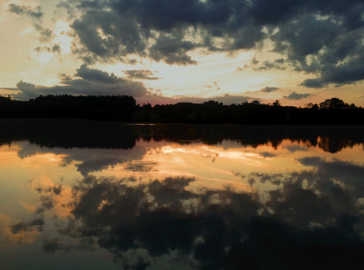 Spokojna tafla jeziora odbija dramatyczne chmury oświetlone zachodzącym słońcem, tworząc symetryczny krajobraz z ciemnym pasem lasu na horyzoncie.