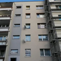 Wielopiętrowy budynek z częściowo odnowioną elewacją, widoczne rusztowania i fragmenty pomalowane na szaro, okna z firankami i anteny satelitarne.
