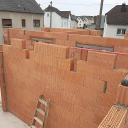 Wznoszenie ściany z czerwonych bloczków ceramicznych na budowie domu, widoczna drewniana drabina oparta o mur i poziomica, w tle zabudowa jednorodzinna.