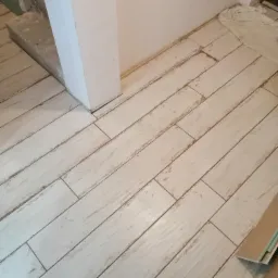 Ułożona podłoga z płytek imitujących drewno w jasnym odcieniu, widoczne fragmenty niedokończonych prac wykończeniowych przy ścianie i schodach, obok leżą płytki i taśma malarska.