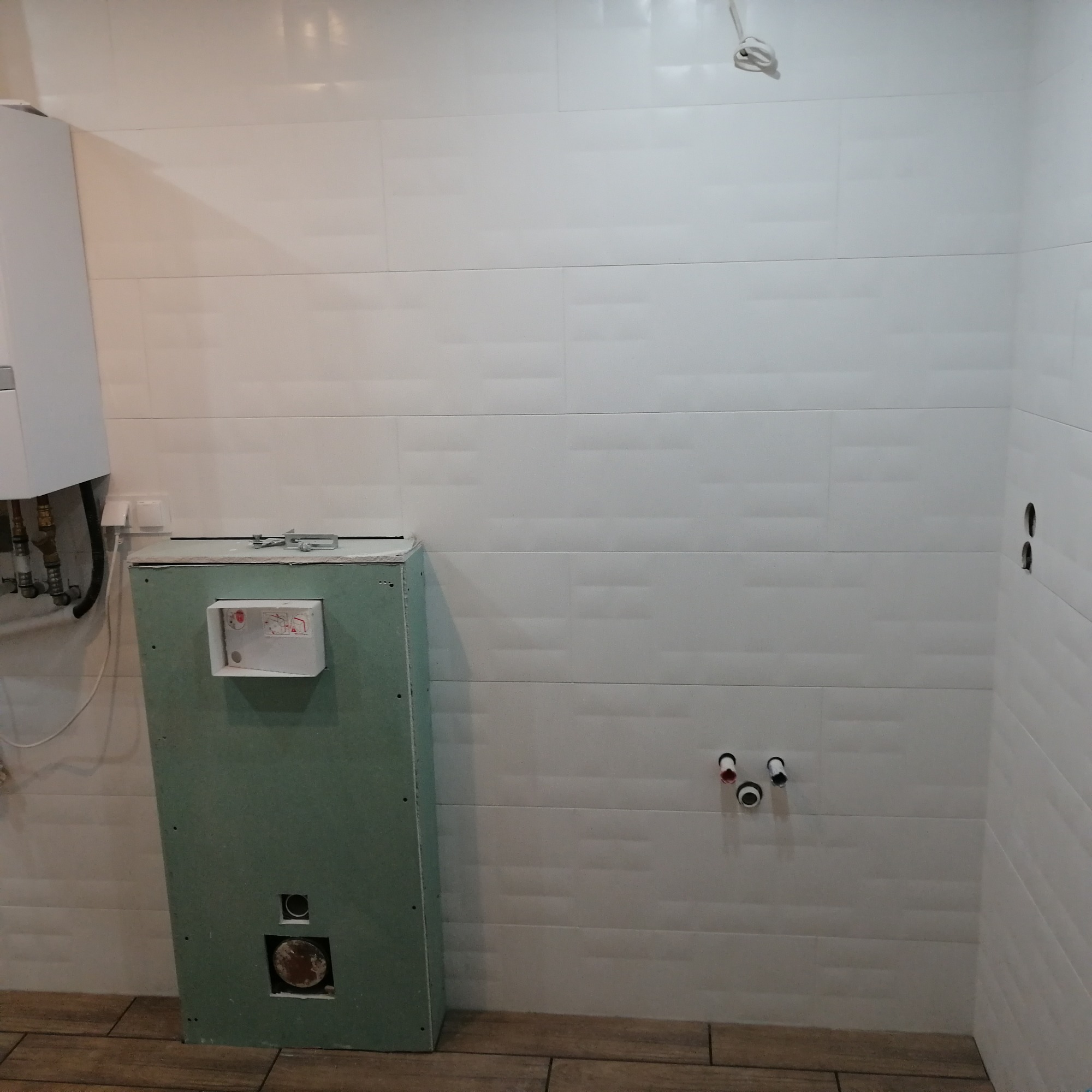 Ściana łazienki w trakcie remontu z widocznymi białymi płytkami, instalacją wodną oraz obudową geberitu z płyty gipsowo-kartonowej, na podłodze ułożone płytki imitujące drewno.