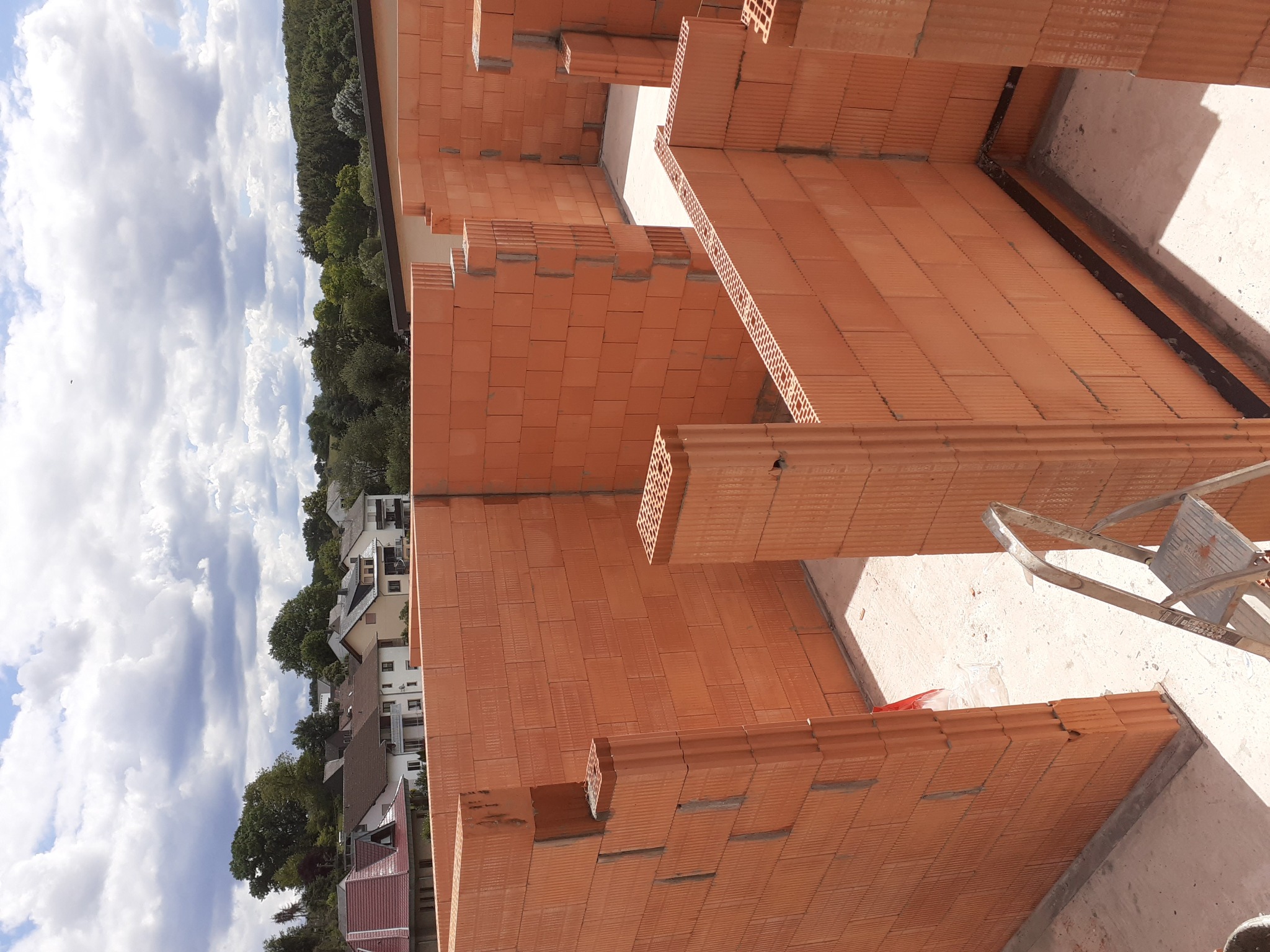 Wznoszone ściany domu z czerwonej ceramiki poryzowanej, widok z góry na budowę, w tle zabudowa jednorodzinna i błękitne niebo z chmurami.