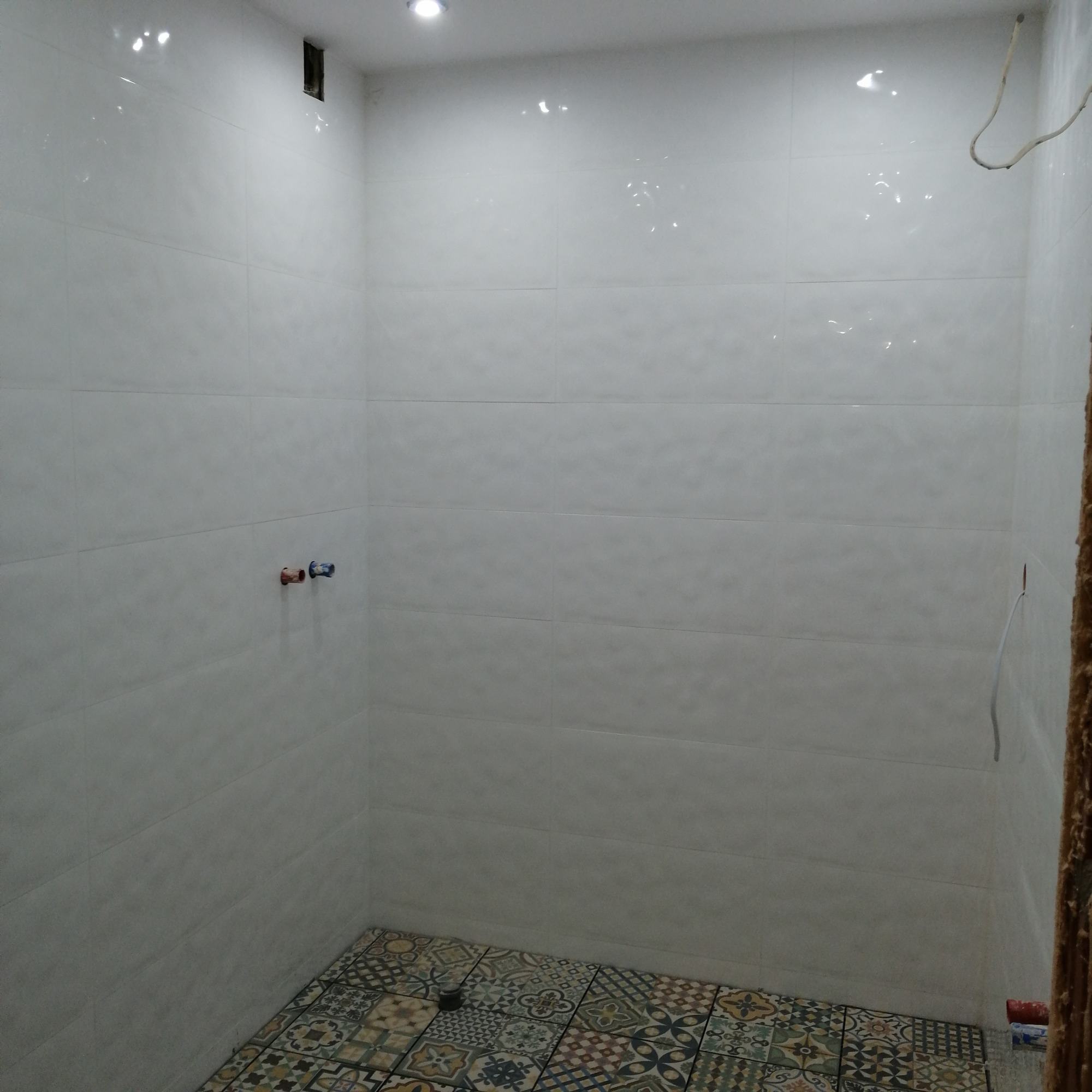 Wnętrze łazienki w trakcie remontu z ułożonymi płytkami podłogowymi w różnokolorowe wzory oraz białymi płytkami ściennymi, widoczne wystające rury instalacji wodnej.