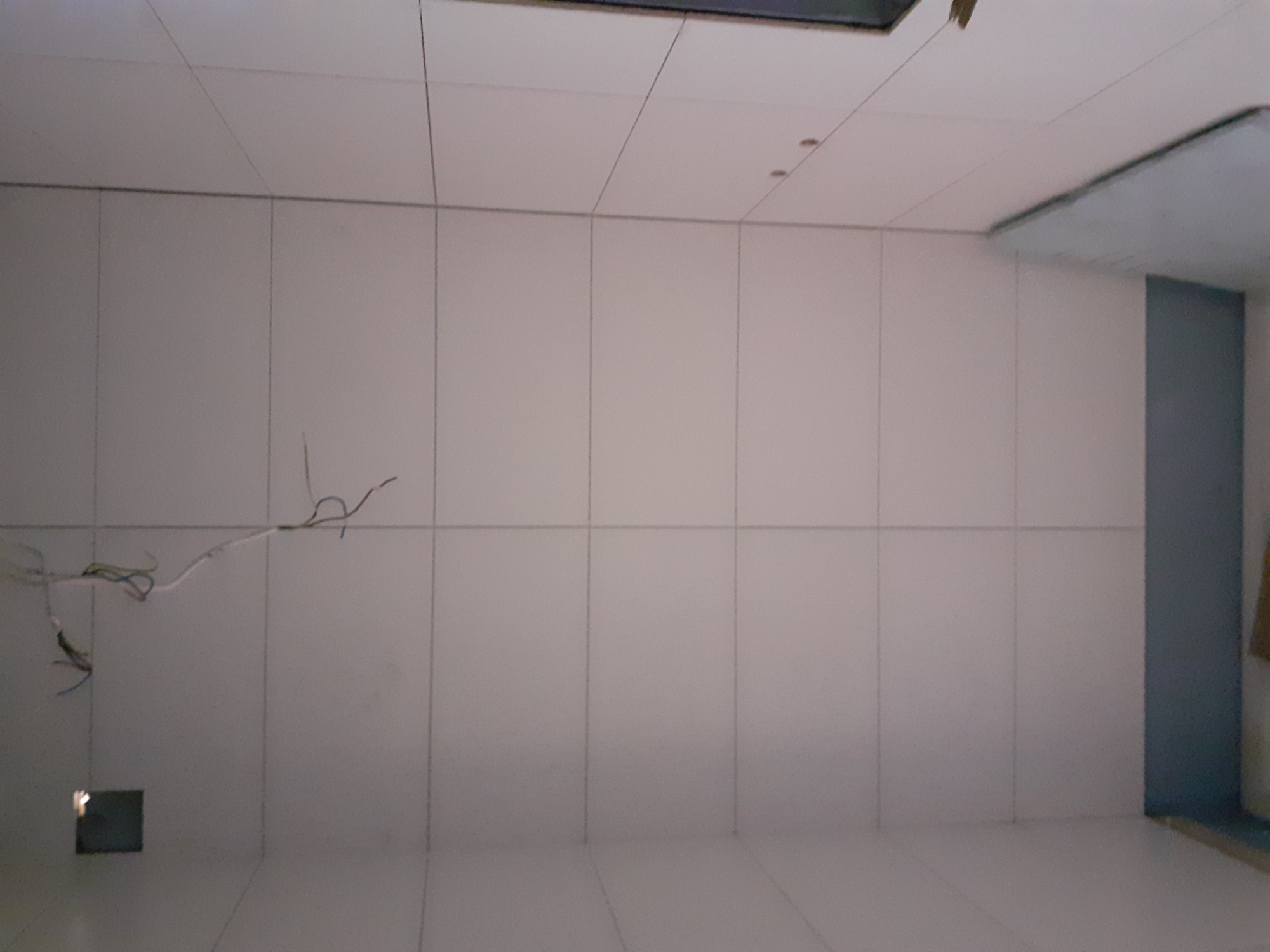 Wnętrze w trakcie prac wykończeniowych: białe płytki ścienne i podłogowe, wystające przewody elektryczne z otworu w ścianie, widoczne elementy oświetlenia sufitowego.