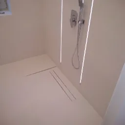 Minimalistyczna łazienka z białymi płytkami, podświetleniem LED i odpływem liniowym. Widoczna bateria prysznicowa z deszczownicą.