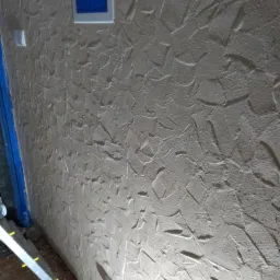 Ściana z dekoracyjnym tynkiem strukturalnym w odcieniach beżu, widoczny fragment okna z niebieską szybą i białą ramą oraz biały włącznik światła, w tle drabina.