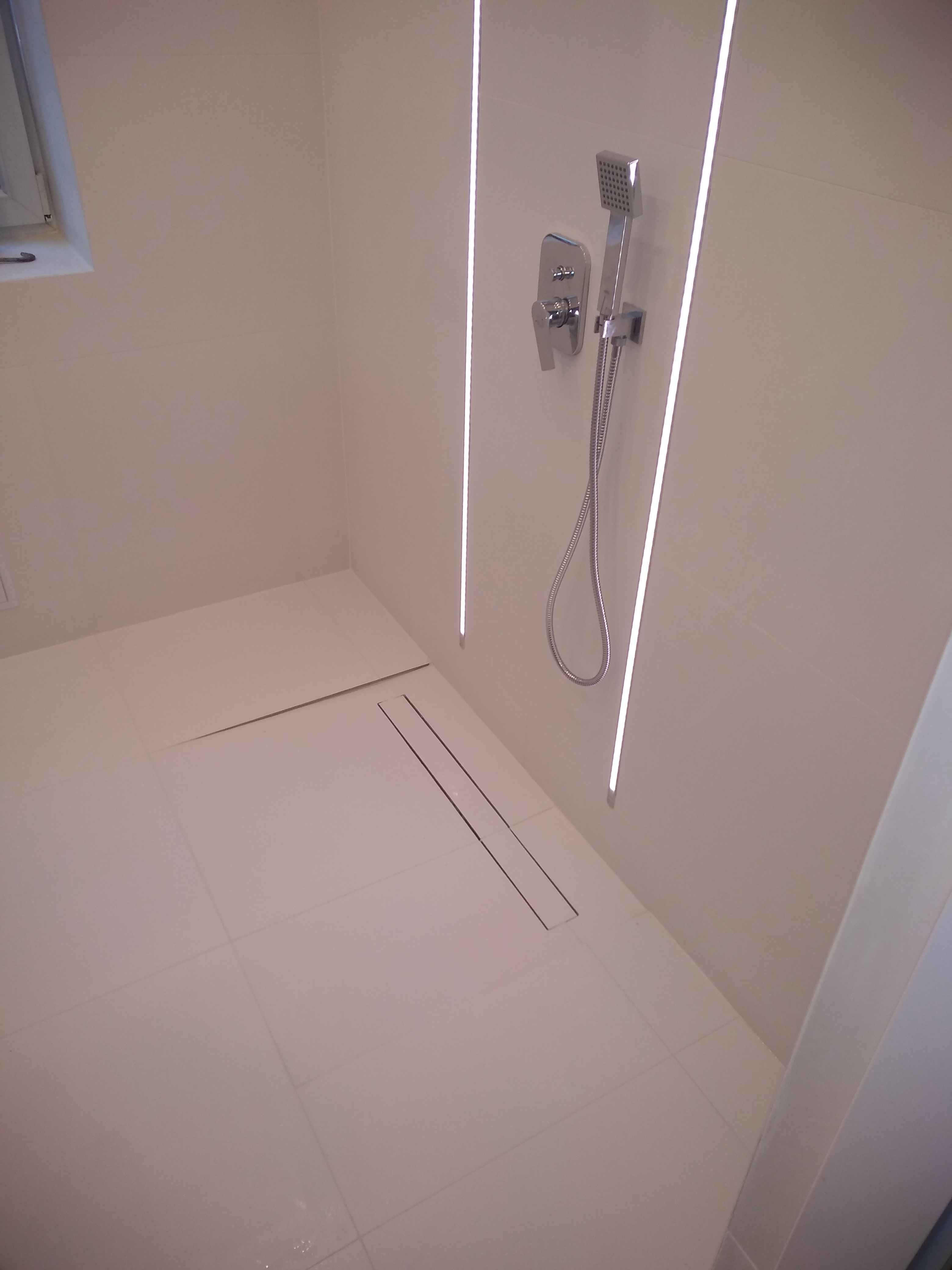 Minimalistyczna łazienka z białymi płytkami, podświetleniem LED i odpływem liniowym. Widoczna bateria prysznicowa z deszczownicą.