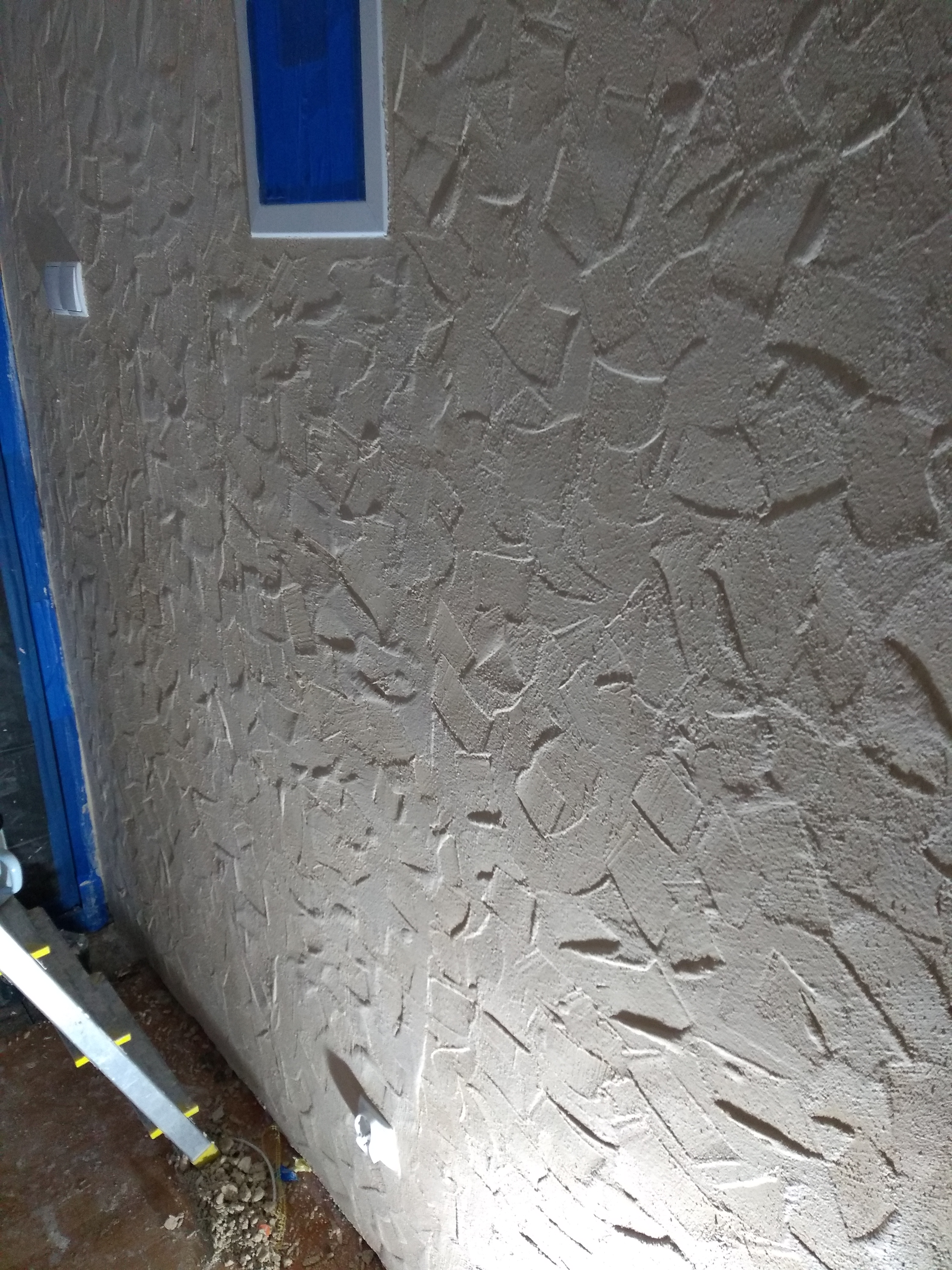 Ściana z dekoracyjnym tynkiem strukturalnym w odcieniach beżu, widoczny fragment okna z niebieską szybą i białą ramą oraz biały włącznik światła, w tle drabina.