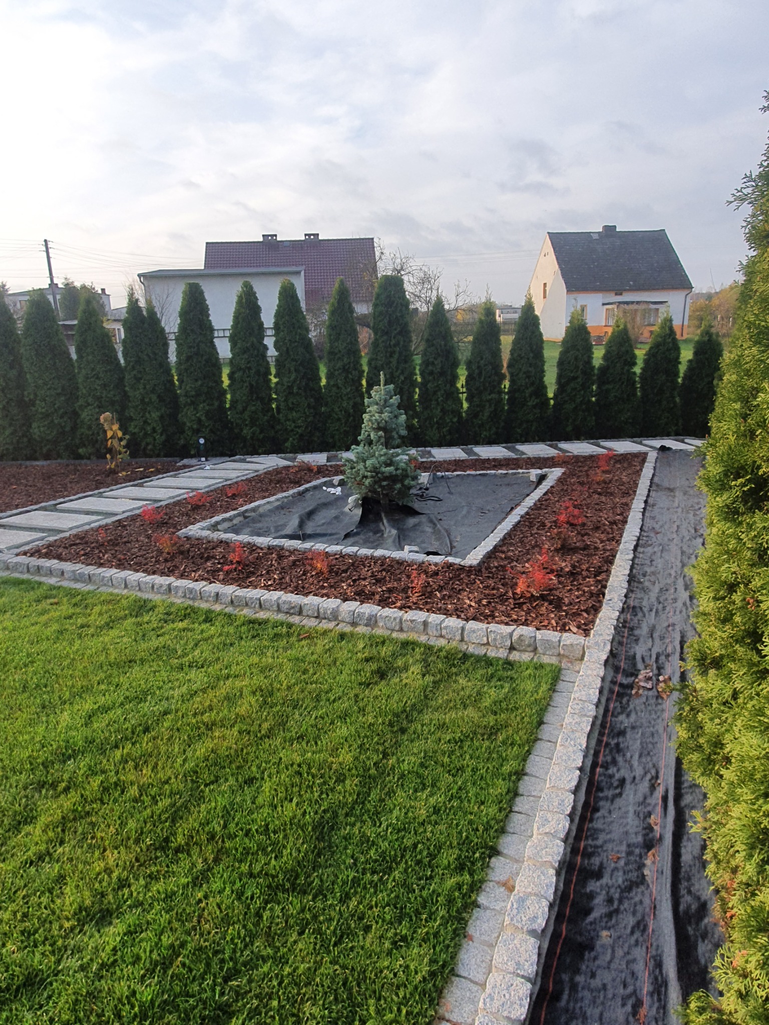 Ogród z równo przystrzyżonym trawnikiem, obrzeżami z kostki granitowej, wyściółką z kory i ścieżką z płyt kamiennych, w tle rząd tuj i zabudowa jednorodzinna.