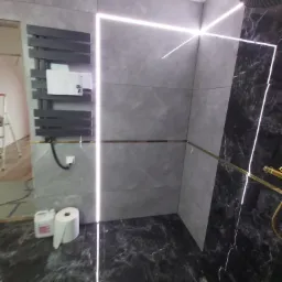 Szare i czarne marmurowe płytki w nowoczesnej łazience z podświetlanym lustrem, grzejnikiem drabinkowym i złotą deszczownicą. Widoczny fragment drabiny malarskiej i rolka papieru toaletowego.