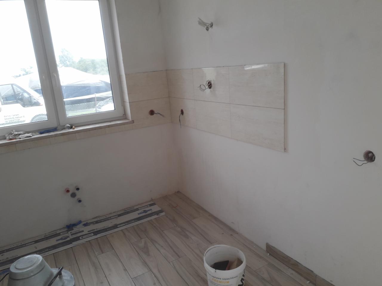 Wnętrze w trakcie remontu: częściowo wyłożone płytki na ścianie, ułożona podłoga z paneli imitujących drewno, widoczne otwory instalacyjne i wiadro z narzędziami.