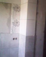 Fragment ściany w trakcie kładzenia płytek. Widoczne otwory na instalację hydrauliczną i częściowo ułożone płytki z motywem kwiatowym, sąsiadujące z pionowym narożnikiem wykończonym białymi płytkami.