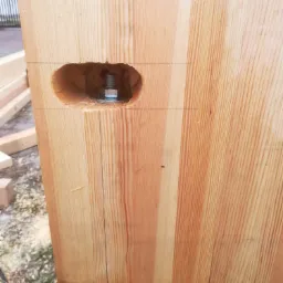 Fragment drewnianej konstrukcji z widocznym otworem na śrubę i podkładką, ukazujący detal połączenia elementów. Widoczne linie pomocnicze naniesione ołówkiem.