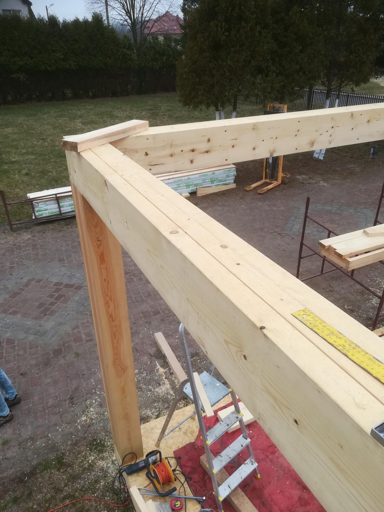 Wznoszenie drewnianej konstrukcji; widoczny fragment szkieletu budynku z jasnego drewna, drabina, narzędzia i materiały budowlane w tle zielony trawnik i drzewa.