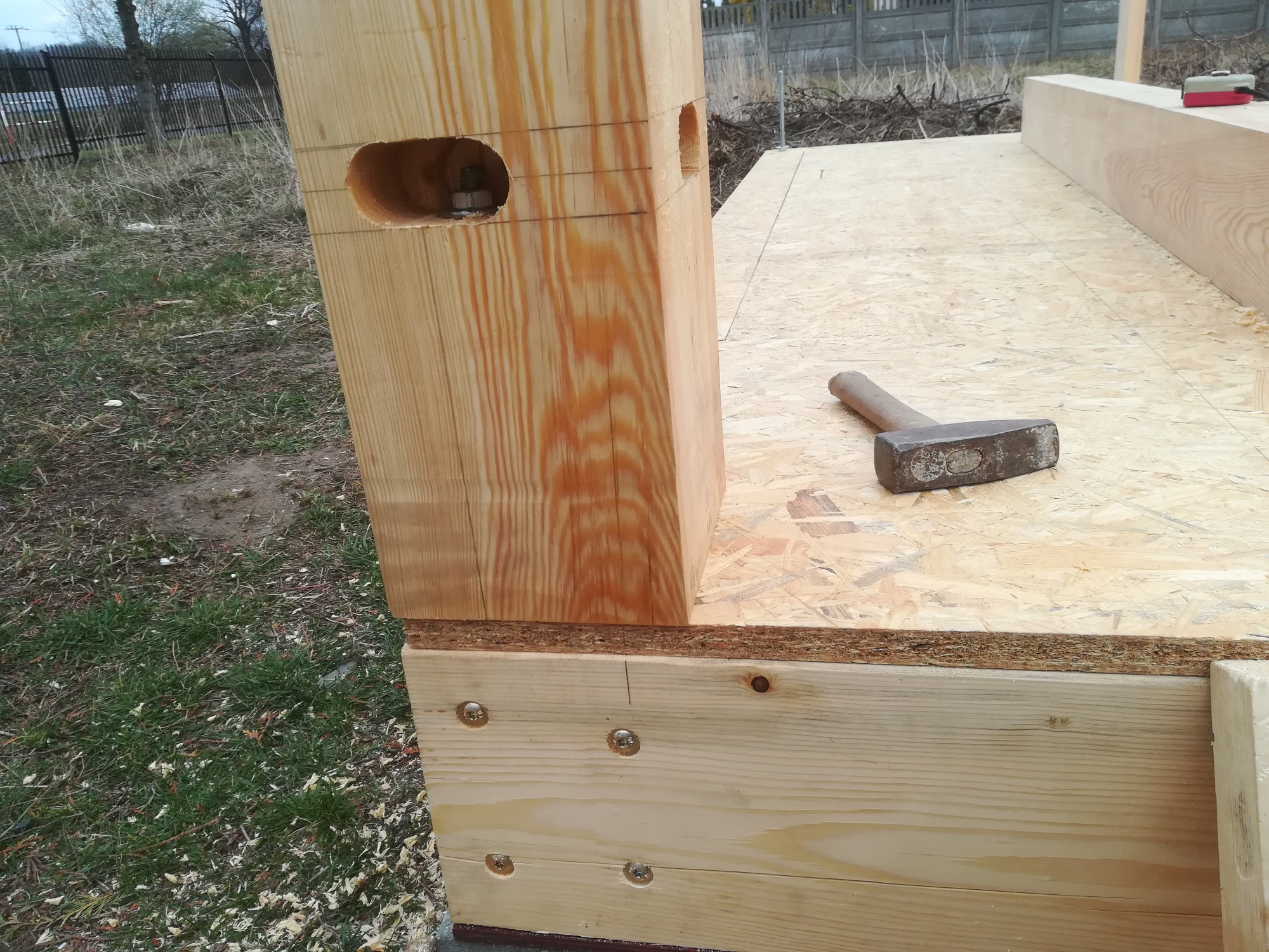 Fragment konstrukcji drewnianej w trakcie montażu, widoczny słup z wywierconymi otworami, połączony śrubami z podstawą, na podłodze leży młotek.