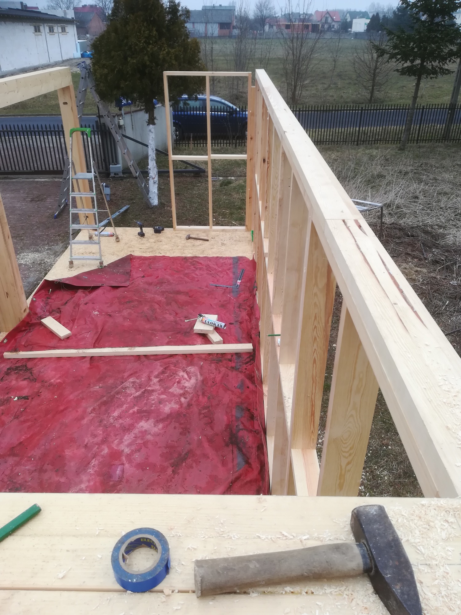 Wznoszenie drewnianej konstrukcji szkieletowej domu: widok z góry na fragment ściany z belek, narzędzia (młotek, taśma, ołówek) oraz plandekę ochronną na podłożu.