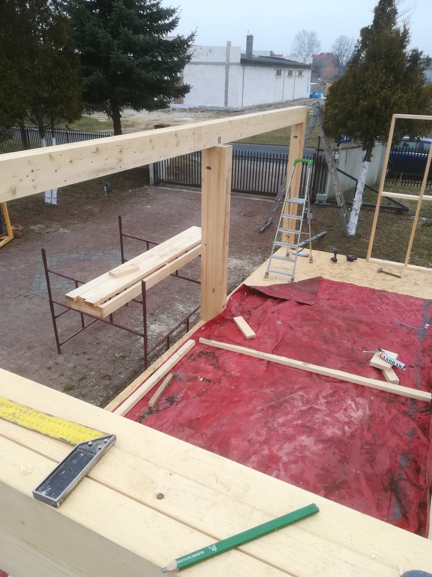 Wznoszenie drewnianej konstrukcji szkieletowej budynku w trakcie budowy, widok z góry, z widocznymi elementami rusztowania, drabiny i narzędzi, zabezpieczona podłoga czerwoną folią.