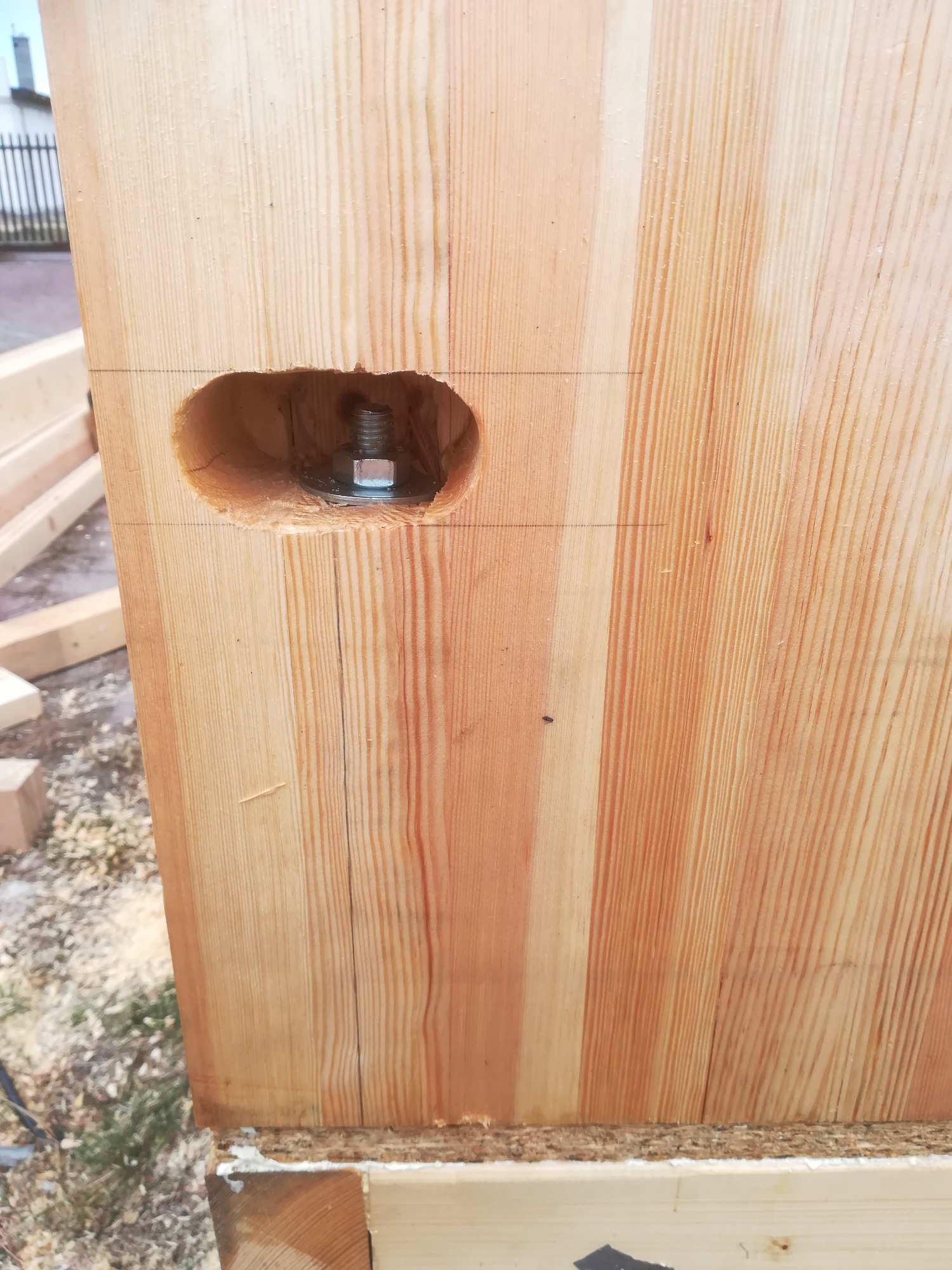 Fragment drewnianej konstrukcji z widocznym otworem na śrubę i podkładką, ukazujący detal połączenia elementów. Widoczne linie pomocnicze naniesione ołówkiem.