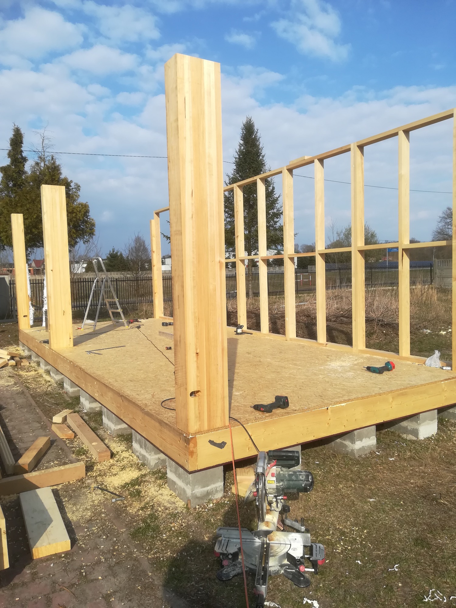 Wznoszenie konstrukcji z drewna na fundamencie betonowym, widoczne narzędzia i elementy konstrukcyjne, w tle błękitne niebo z chmurami.