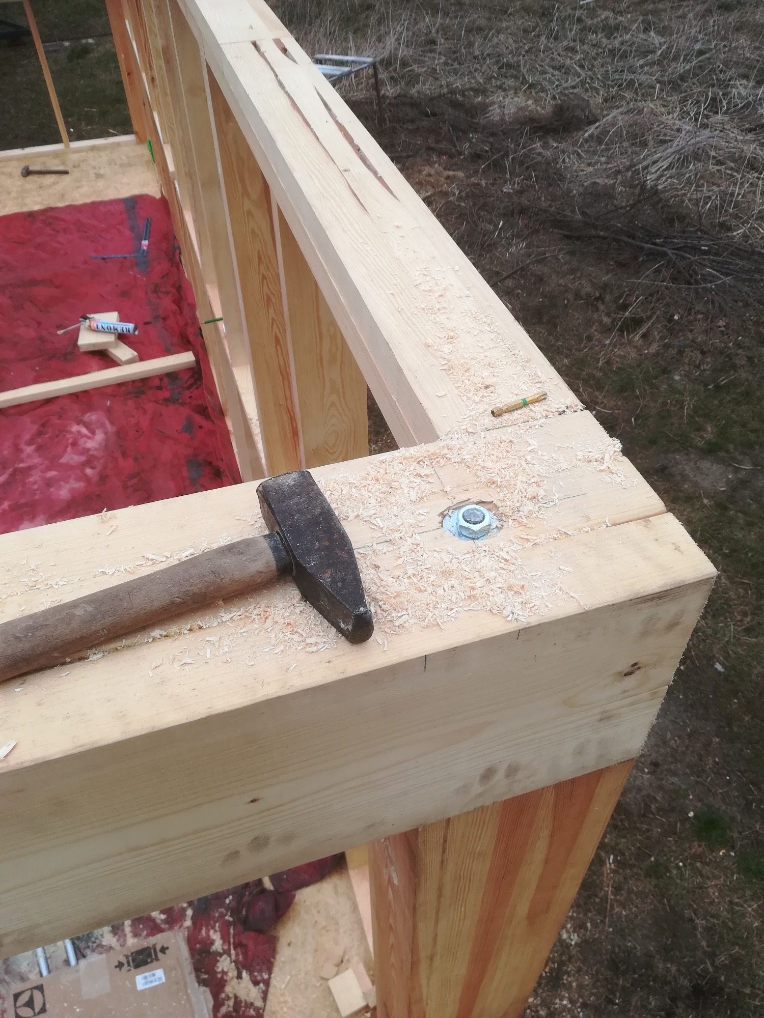 Fragment drewnianej konstrukcji w trakcie montażu, widoczny młotek leżący na belce oraz ślady wiercenia i wióry, w tle czerwona plandeka i fragment krajobrazu.