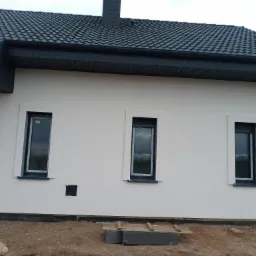 Nowa elewacja budynku jednorodzinnego w Kołobrzegu, z trzema oknami i widocznym fragmentem dachu z ciemnej dachówki. Świeżo położony jasny tynk elewacyjny. Teren wokół budynku jeszcze...