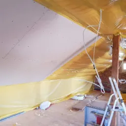 Poddasze w trakcie prac: zamontowana płyta kartonowo-gipsowa na skosie, zabezpieczone folią ściany, widoczne instalacje elektryczne i drabina.