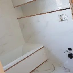 Biała łazienka z wanną, płytkami imitującymi marmur i złotymi listwami dekoracyjnymi, widoczne przyłącza wodne i zabudowany stelaż podtynkowy WC.