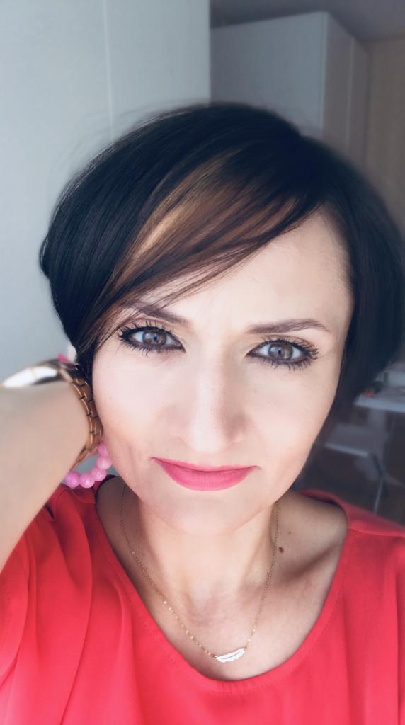 Portret kobiety z krótkimi ciemnymi włosami z pasemkiem w kolorze miodowym, w czerwonej bluzce i delikatnej złotej biżuterii z motywem pióra.