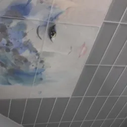 Ukośne ujęcie ściany wyłożonej szarymi kafelkami, z fragmentem dekoracyjnej mozaiki przedstawiającej oko i kwiatowe motywy w odcieniach błękitu i szarości.