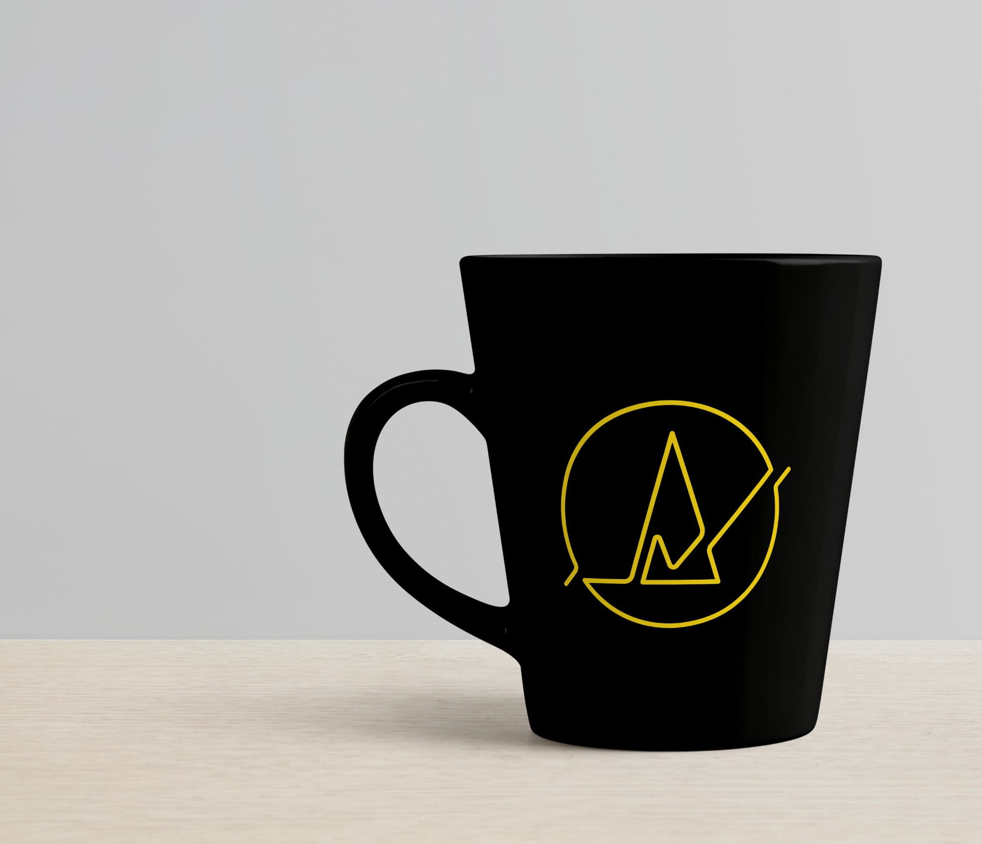 Czarny kubek z żółtym, minimalistycznym logo przedstawiającym stylizowany szczyt góry w okręgu, stojący na jasnym drewnianym blacie na tle szarej ściany.