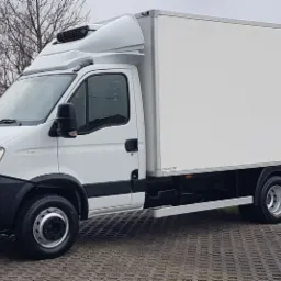 Biały samochód dostawczy Iveco z chłodnią, zaparkowany na kostce brukowej, widok z boku na tle drzew bez liści.
