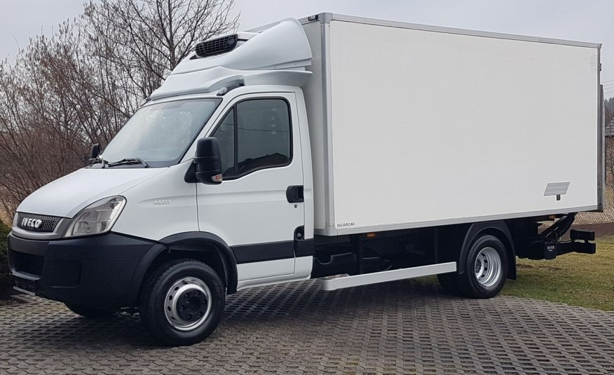 Biały samochód dostawczy Iveco z chłodnią, zaparkowany na kostce brukowej, widok z boku na tle drzew bez liści.