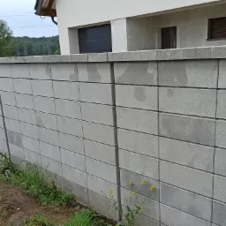 Nowoczesne ogrodzenie murowane z szarych bloczków modułowych, widok na dom z garażem i zieleń w tle.