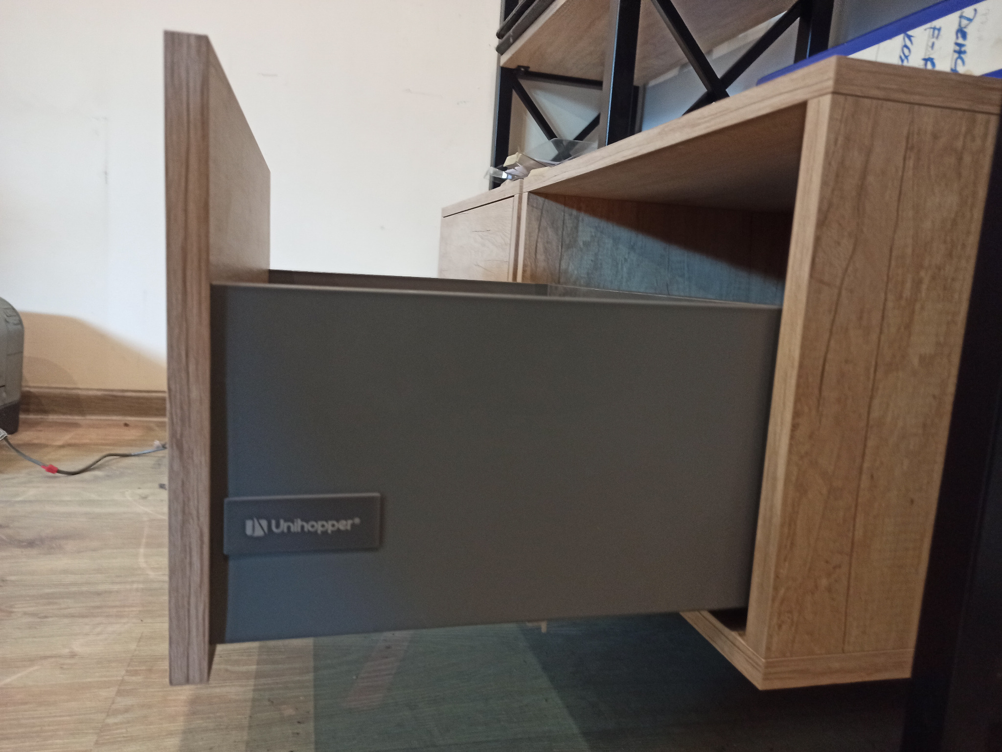 Wysuwana szuflada z szarą, matową powierzchnią i widocznym logo Unihoper, zamontowana w szafce z drewnopodobną okleiną i czarnymi metalowymi elementami konstrukcyjnymi.