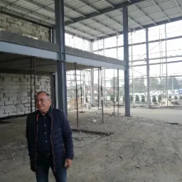 Mężczyzna w kurtce na terenie budowy hali z widoczną konstrukcją stalową, ścianami z bloczków i dużymi oknami.