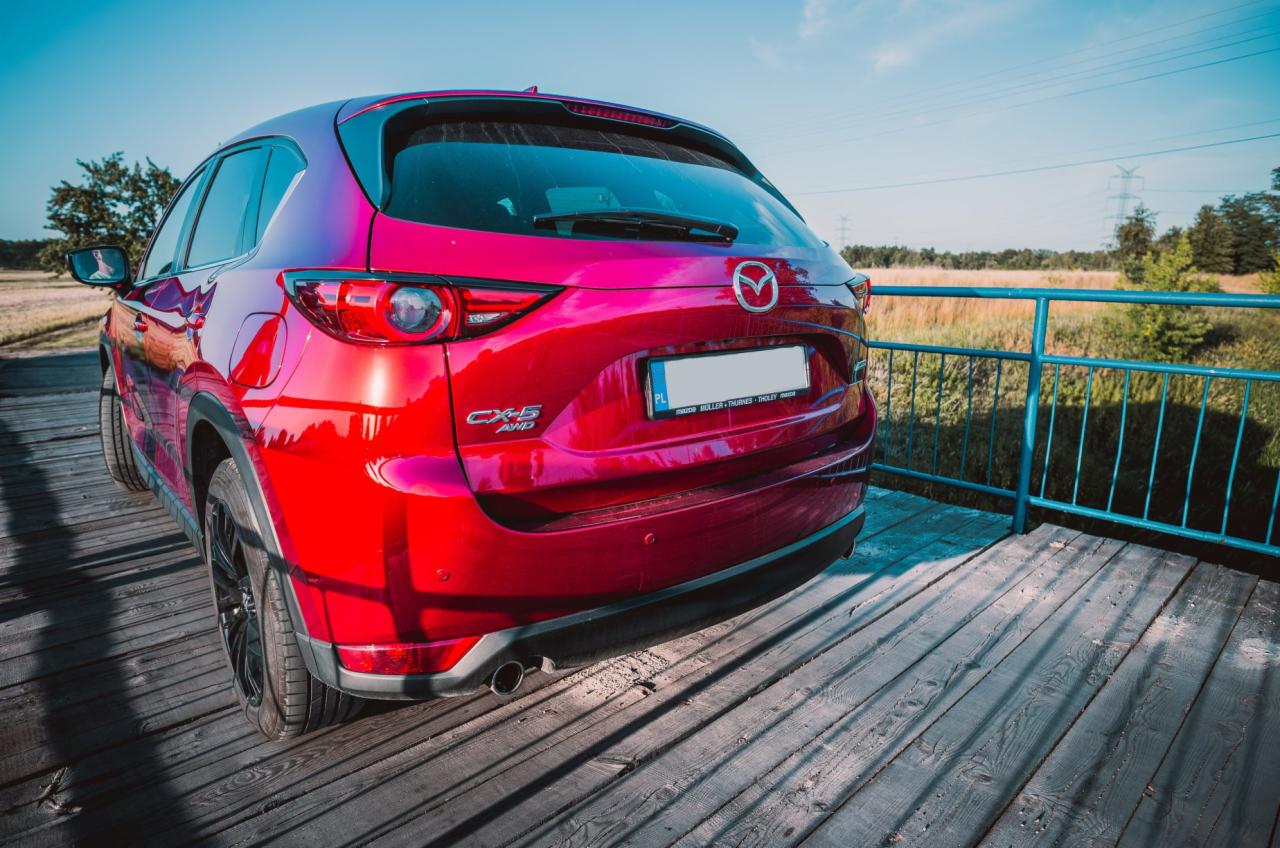 Czerwony samochód Mazda CX-5 AWD, widziany z tyłu, zaparkowany na drewnianym pomoście z niebieską barierką, z polską tablicą rejestracyjną, w tle łąka i linia wysokiego napięcia.
