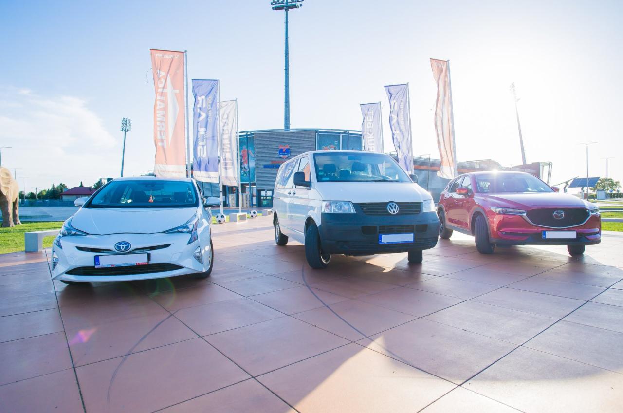 Trzy różne modele samochodów (Toyota Prius, Volkswagen Transporter, Mazda CX-5) zaparkowane przed budynkiem z flagami w słoneczny dzień.