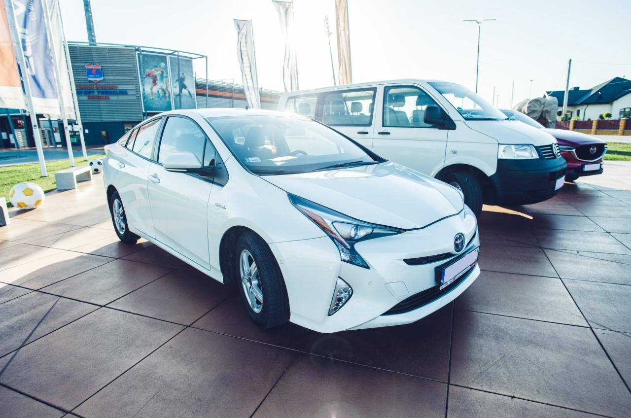 Biała Toyota Prius zaparkowana na placu przed budynkiem, obok biały van i czerwony samochód osobowy.