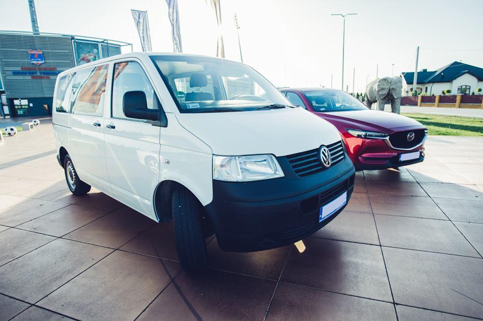 Biały van Volkswagen i czerwony samochód osobowy Mazda zaparkowane na placu z płyt, w tle budynek z logo i rzeźba słonia.