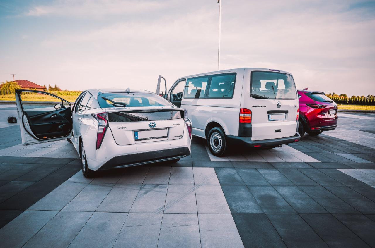 Trzy samochody: biała Toyota Prius z otwartymi drzwiami, biały Volkswagen Transporter i czerwona Mazda CX-30 stojące na parkingu z kostki brukowej.