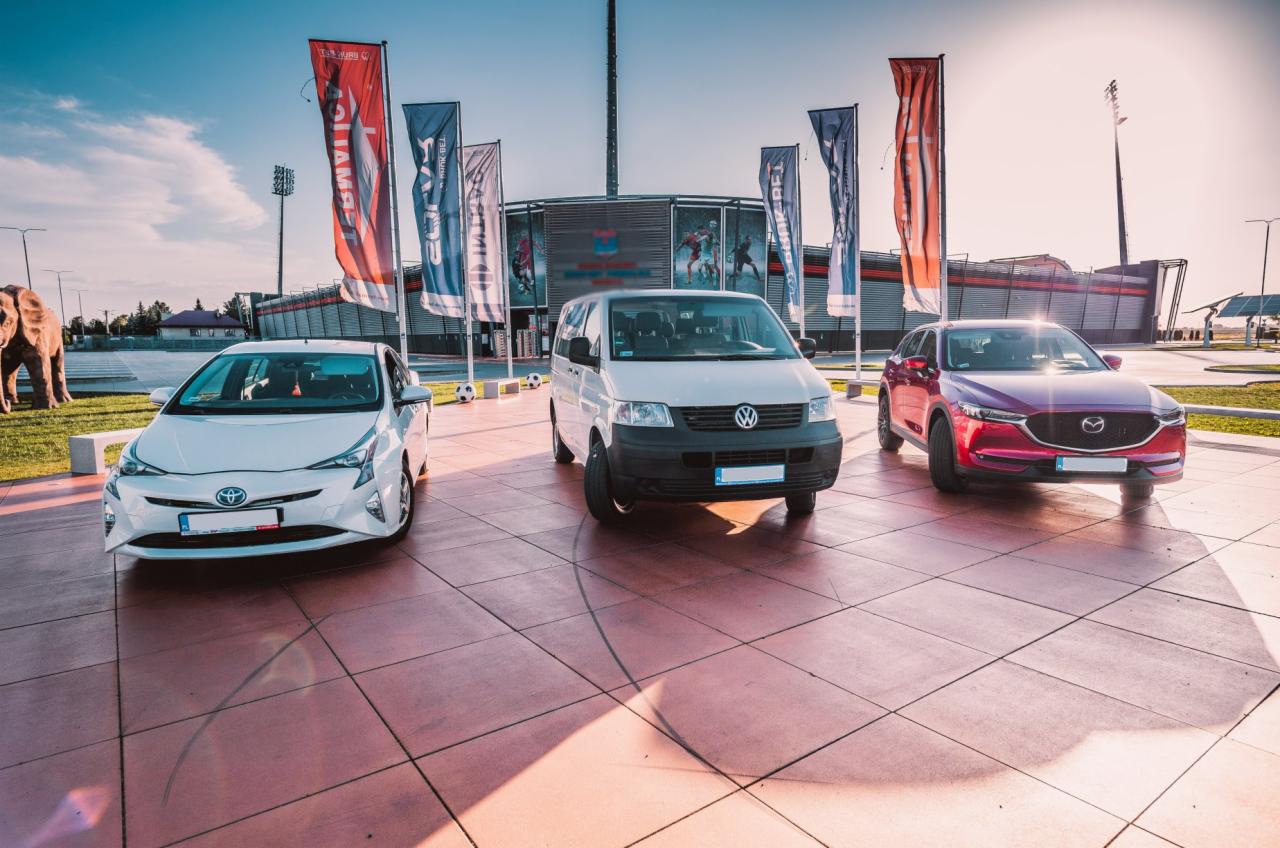 Trzy samochody (biały Toyota Prius, biały Volkswagen Transporter, bordowy Mazda CX-5) zaparkowane na placu przed stadionem piłkarskim z powiewającymi flagami i rzeźbą słonia w tle.