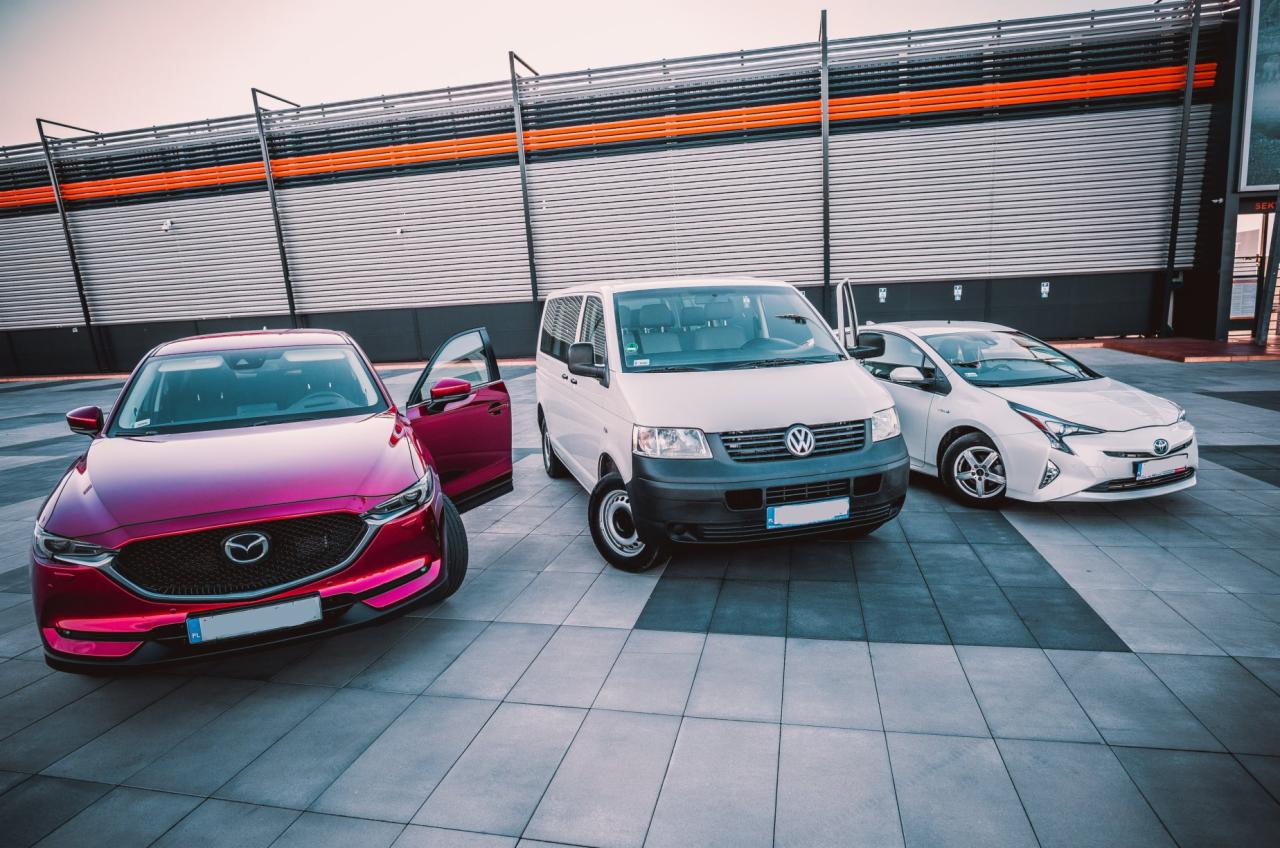 Trzy samochody: bordowa Mazda z otwartymi drzwiami, biały Volkswagen Transporter i biała Toyota Prius, stojące na placu z szarej kostki, na tle budynku z metalową fasadą i pomarańczowymi akcentami.