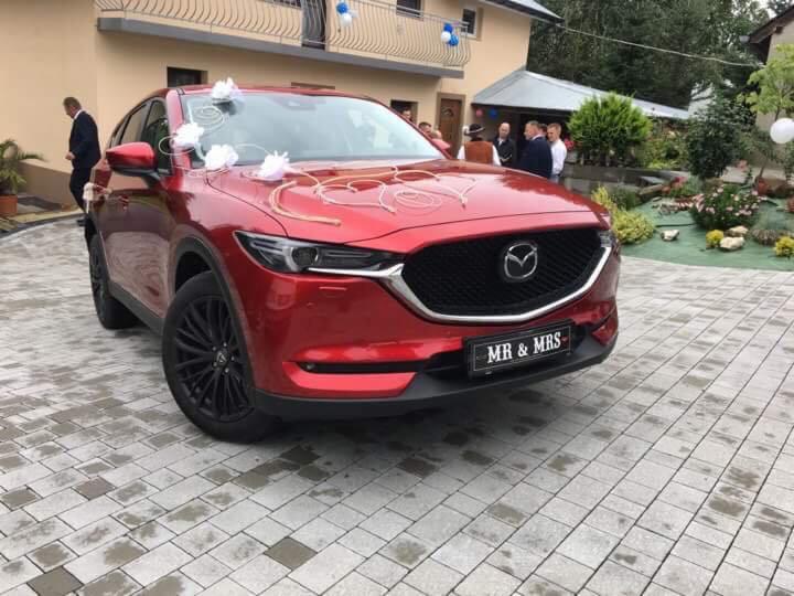 Czerwona Mazda CX-5 z czarnymi felgami, udekorowana białymi kokardami i napisem 'MR & MRS' na tablicy rejestracyjnej, przygotowana do ślubu przed budynkiem z balkonem.