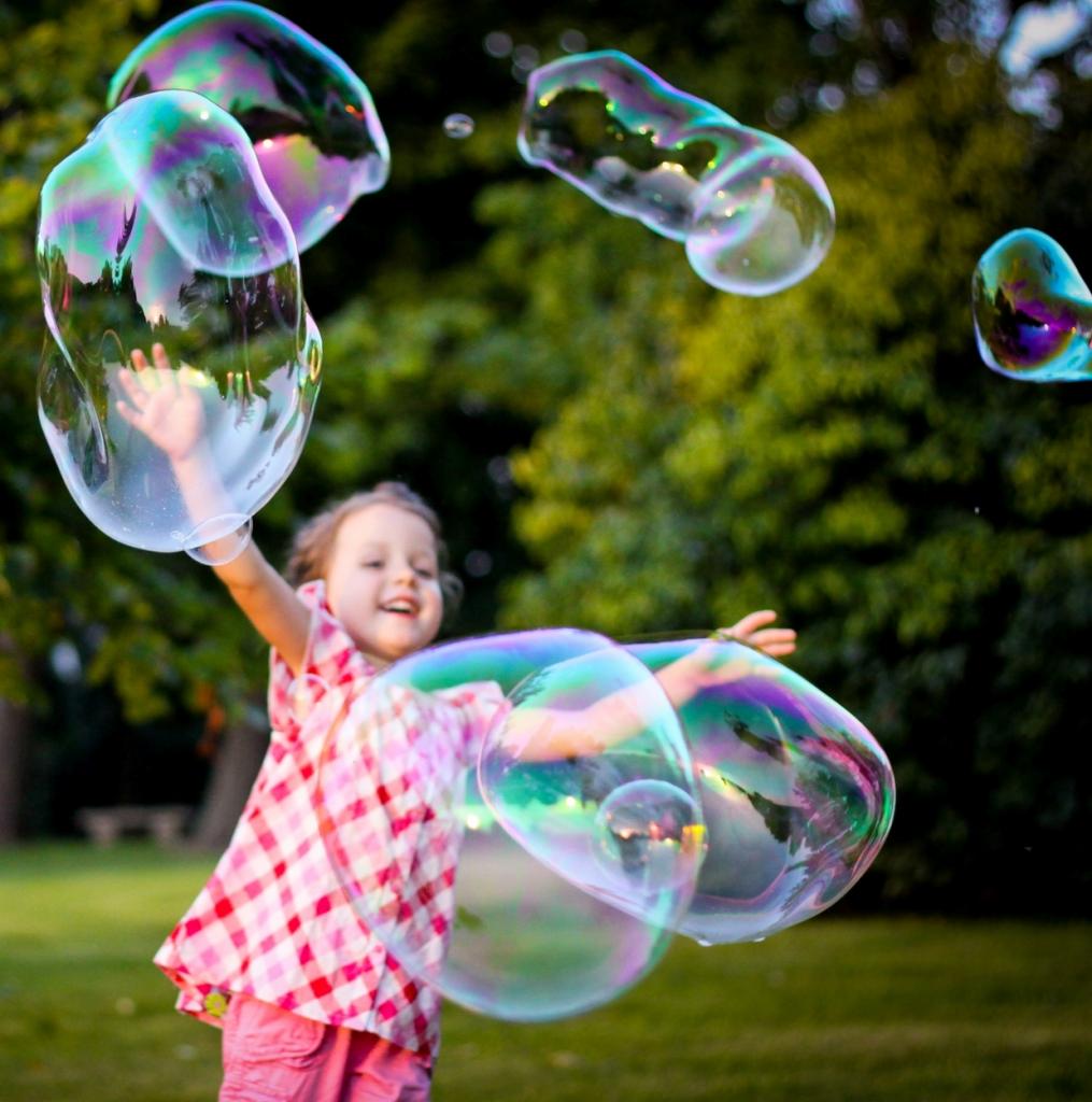 Dziewczynka w różowej koszuli w kratę bawi się dużymi, tęczowymi bańkami mydlanymi na tle zielonego ogrodu.