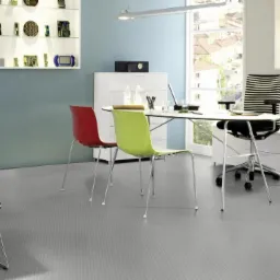 Nowoczesne biuro z szarą podłogą o delikatnym wzorze, krzesłem w pepitkę i kolorowymi krzesłami przy białym biurku.