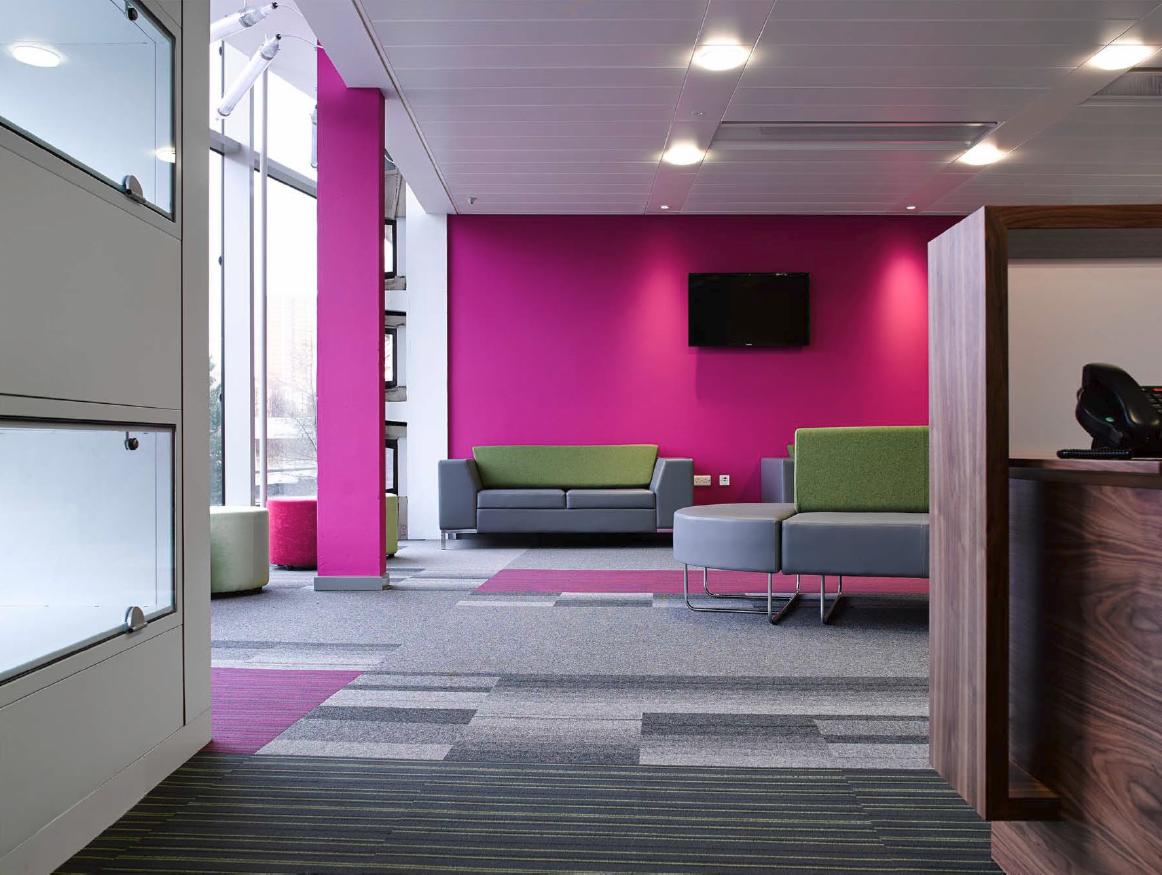 Nowoczesne biuro z wykładziną dywanową w geometryczne wzory, fuksjowa ściana, szare sofy z zielonymi akcentami i recepcja z ciemnego drewna.